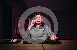 Handsome happy guy in a headset around his neck sits at a table at a computer in a room with a purple light and calls on the phone