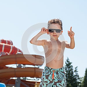 Handsome happy little caucasian boy in sunglasses showing number one finger gesture in summer water park during vacation