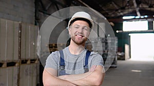 Handsome and happy professional worker in hat charmingly smiling on camera. In the background big warehouse