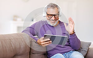 Handsome happy senior man waving by hand while talking online on digital tablet with family