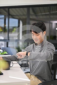 Handsome Asian male developer or office worker checking time on his smartwatch at his desk