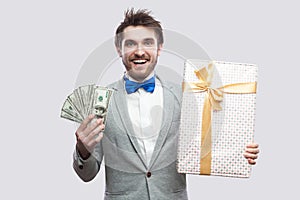 Handsome happy young bearded man in gray suit and blue bow tie standing and holding gift box with yellow bow and many cash dollars