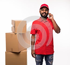 Handsome happy young delivery man talking by mobile phone in red cap standing with parcel post boxes isolated over white