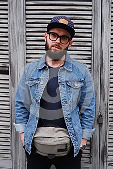 Handsome hipster guy in a cap and glasses with a beard in a blue denim jacket and a fanny pack