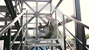 Handsome hipster guy using a smartphone sitting on the iron stairs of an industrial building. Beautiful young man using