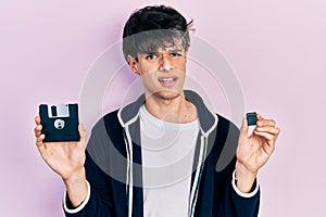 Handsome hipster young man holding floppy disk and sdxc card clueless and confused expression