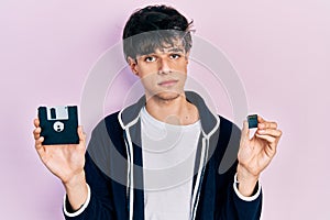 Handsome hipster young man holding floppy disk and sdxc card relaxed with serious expression on face