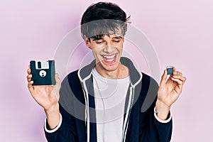 Handsome hipster young man holding floppy disk and sdxc card smiling and laughing hard out loud because funny crazy joke