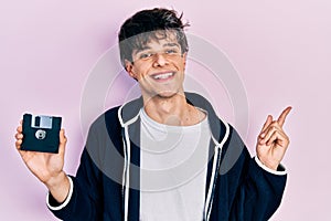 Handsome hipster young man holding floppy disk smiling happy pointing with hand and finger to the side