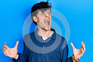 Handsome hipster young man stranding over blue background crazy and mad shouting and yelling with aggressive expression and arms