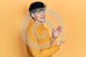 Handsome hipster young man wearing casual yellow sweater inviting to enter smiling natural with open hand