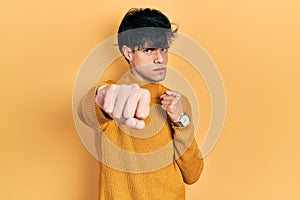 Handsome hipster young man wearing casual yellow sweater punching fist to fight, aggressive and angry attack, threat and violence