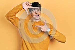 Handsome hipster young man wearing casual yellow sweater smiling making frame with hands and fingers with happy face