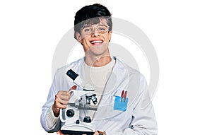 Handsome hipster young man wearing lab coat holding microscope smiling with a happy and cool smile on face