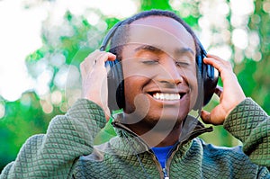 Handsome hispanic black man wearing green sweater in outdoors park area, hands holding headphones covering ears and