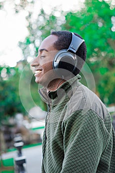 Handsome hispanic black man wearing green sweater in outdoors park area, headphones on covering ears and smiling