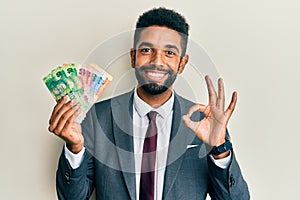 Handsome hispanic business man with beard holding south african rand banknotes doing ok sign with fingers, smiling friendly