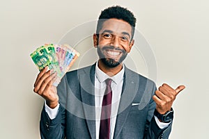 Handsome hispanic business man with beard holding south african rand banknotes pointing thumb up to the side smiling happy with