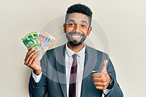 Handsome hispanic business man with beard holding south african rand banknotes smiling happy and positive, thumb up doing