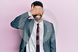 Handsome hispanic business man with beard wearing business suit and tie smiling and laughing with hand on face covering eyes for