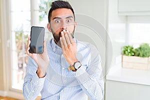 Handsome hispanic business man showing smartphone screen cover mouth with hand shocked with shame for mistake, expression of fear,