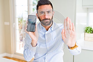 Handsome hispanic business man showing smartphone screen with open hand doing stop sign with serious and confident expression,