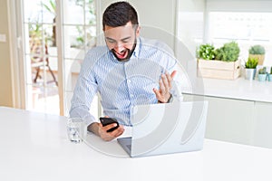 Handsome hispanic business man using laptop and smartphone very happy and excited, winner expression celebrating victory screaming