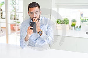 Handsome hispanic business man using smartphone cover mouth with hand shocked with shame for mistake, expression of fear, scared