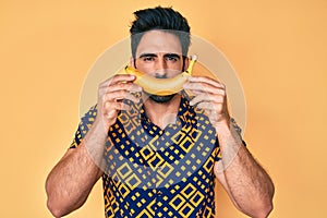 Handsome hispanic man with beard holding banana like funny smile relaxed with serious expression on face