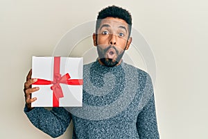 Handsome hispanic man with beard holding gift scared and amazed with open mouth for surprise, disbelief face