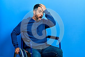 Handsome hispanic man with beard sitting on wheelchair surprised with hand on head for mistake, remember error