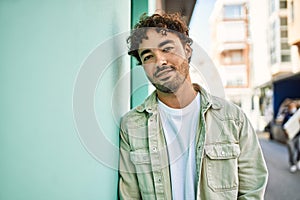 Handsome hispanic man with beard smiling happy outdoors on a sunny day leaning on a wall