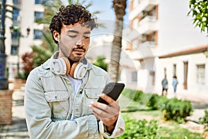 Handsome hispanic man with beard smiling happy outdoors on a sunny day wearing headphones listening to music with smartphone