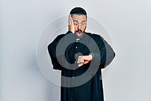 Handsome hispanic man with beard wearing catholic priest robe looking at the watch time worried, afraid of getting late