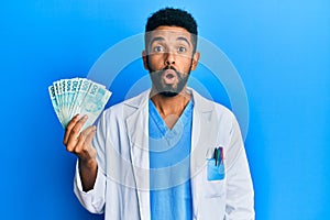 Handsome hispanic man with beard wearing doctor uniform holding 100 brazilian reals scared and amazed with open mouth for