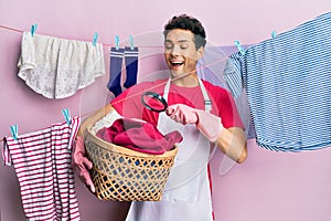 Handsome hispanic man doing laundry holding wicker basket looking for a stain celebrating crazy and amazed for success with open