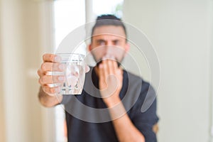 Handsome hispanic man drinking a fresh glass of water cover mouth with hand shocked with shame for mistake, expression of fear,