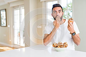 Handsome hispanic man eating chocolate chips muffin cover mouth with hand shocked with shame for mistake, expression of fear,