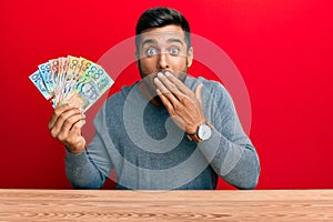 Handsome hispanic man holding australian dollars covering mouth with hand, shocked and afraid for mistake