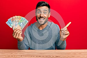 Handsome hispanic man holding australian dollars smiling happy pointing with hand and finger to the side