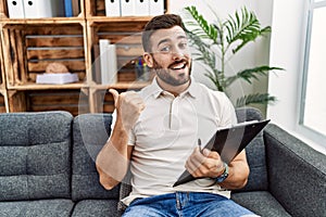 Handsome hispanic man holding clipboard working at psychology clinic pointing to the back behind with hand and thumbs up, smiling