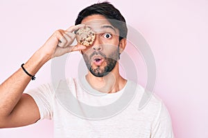Handsome hispanic man holding cookie scared and amazed with open mouth for surprise, disbelief face