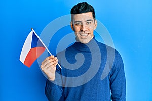 Handsome hispanic man holding czech republic flag looking positive and happy standing and smiling with a confident smile showing