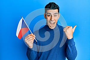 Handsome hispanic man holding czech republic flag pointing thumb up to the side smiling happy with open mouth