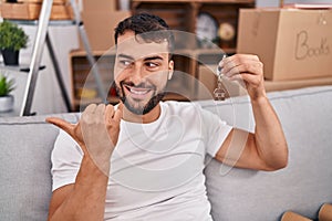 Handsome hispanic man holding keys of new home pointing thumb up to the side smiling happy with open mouth