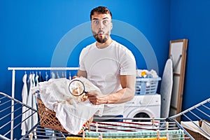 Handsome hispanic man holding magnifying glass looking for stain at clothes making fish face with mouth and squinting eyes, crazy