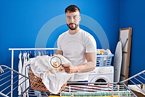 Handsome hispanic man holding magnifying glass looking for stain at clothes relaxed with serious expression on face