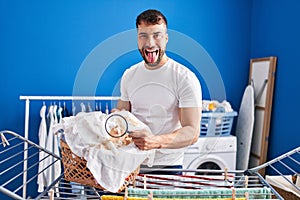 Handsome hispanic man holding magnifying glass looking for stain at clothes sticking tongue out happy with funny expression