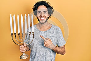 Handsome hispanic man holding menorah hanukkah jewish candle smiling happy pointing with hand and finger