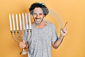 Handsome hispanic man holding menorah hanukkah jewish candle smiling happy pointing with hand and finger to the side